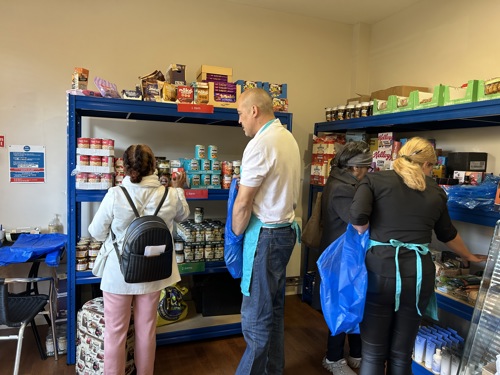 Four people standing with their backs to the cameral choosing items of food from blue shelving staked high with tins and packets of long-life food. Tow of the people have Peabody teal aprons on and are helping the other two choose items of food.