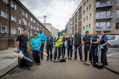 Group of Peabody colleagues dressed in Peabody workwear standing in a semicircle facing the camera holding long-handled brushes, mops, leaf blowers and other cleaning materials.